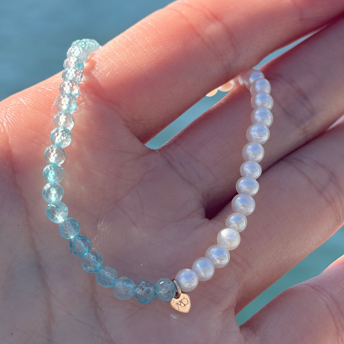 The Mermaid's Den Pearly Me bracelet featuring freshwater pearls and blue gemstones on a hand against a water backdrop.
