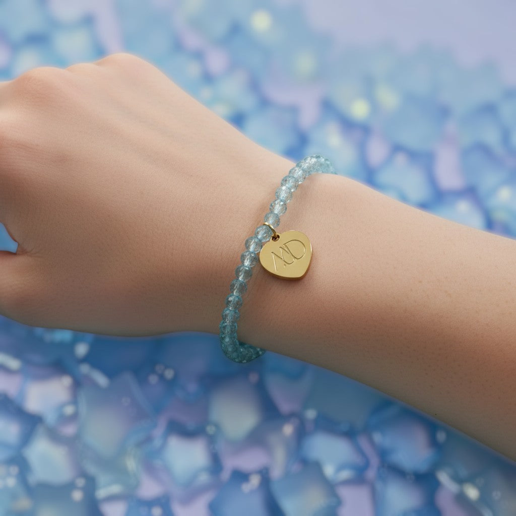 The Mermaid's Den Tweedia aquamarine beaded bracelet with a gold heart charm worn on a wrist against a blue background