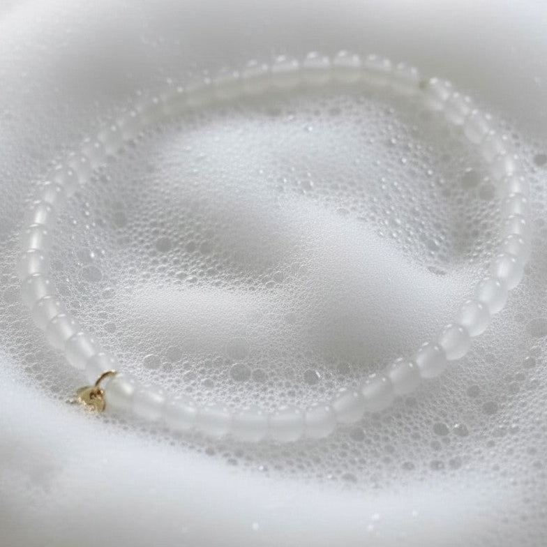 The Mermaid's Den white jade beaded bracelet with a small gold charm resting on soft white soap bubbles.
