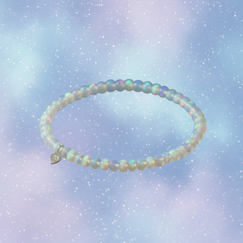 The Mermaid's Den Hyacinthus White Opal beaded bracelet with a small silver heart charm on a dreamy pastel background.